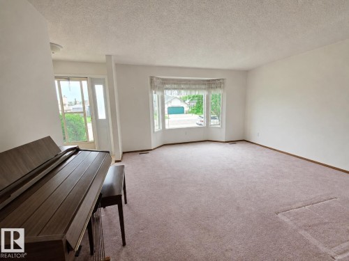 Expansive carpeted living space featuring a prominent bay window, white painted walls, and a front entry door with a sidelight - 4005 55 Avenue, Whitecourt, AB - Indoor Photo Showing Other Room