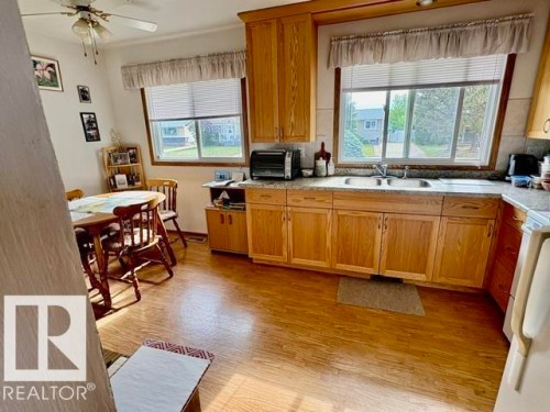 Kitchen featuring wood-finish flooring, wood cabinetry, L-shaped countertop, double basin sink, and a ceiling fan - 5454 46 Street, Vegreville, AB - Indoor Photo Showing Kitchen With Double Sink