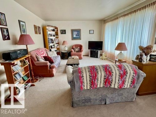 Carpeted living room featuring a wide window with vertical blinds, light-toned walls, and crown molding - 5454 46 Street, Vegreville, AB - Indoor