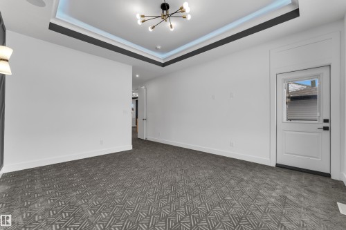 Expansive room featuring patterned carpeting, a tray ceiling with integrated lighting, a contemporary chandelier, white walls, and a white exterior door with a window - 2027 156 Avenue, Edmonton, AB - Indoor Photo Showing Other Room