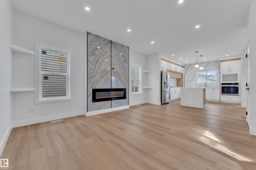 Open-concept living space featuring a modern fireplace with a marble-look surround, light wood-finish flooring, recessed lighting, and a kitchen with an island, built-in appliances, and pendant lighting - 6092 179 Ave Nw, Edmonton, AB - Indoor Photo Showing Living Room With Fireplace