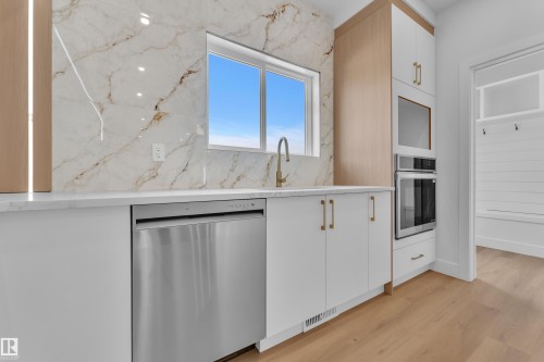 Kitchen area featuring a window, light wood-finish flooring, white cabinetry with gold-tone hardware, a stainless steel dishwasher, and a built-in stainless steel oven - 6092 179 Ave Nw, Edmonton, AB - Indoor Photo Showing Kitchen