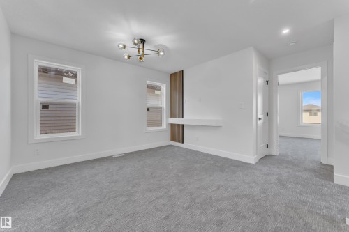 Spacious room featuring light gray carpeting, a modern multi-bulb ceiling fixture, two white-framed windows, and a built-in floating shelf with a wood-finish accent panel - 6092 179 Ave Nw, Edmonton, AB - Indoor Photo Showing Other Room