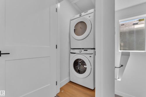 Dedicated laundry area with stacked front-load washer and dryer, hardwood flooring, modern white doors with black hardware, and a nearby window providing natural light - 6092 179 Ave Nw, Edmonton, AB - Indoor Photo Showing Laundry Room
