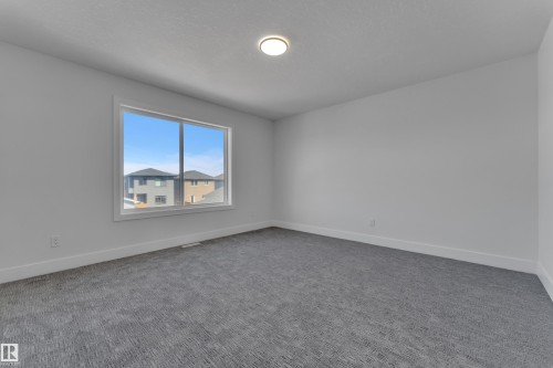 Spacious room featuring a large window with white trim, light gray walls, dark patterned carpeting, and a recessed ceiling light - 6092 179 Ave Nw, Edmonton, AB - Indoor Photo Showing Other Room