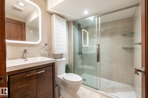 Modern bathroom featuring a wood-finish vanity with an integrated white sink, a sleek wall-mounted mirror, a frameless glass shower enclosure, and a contemporary toilet - 4632 151 Street, Edmonton, AB - Indoor Photo Showing Bathroom