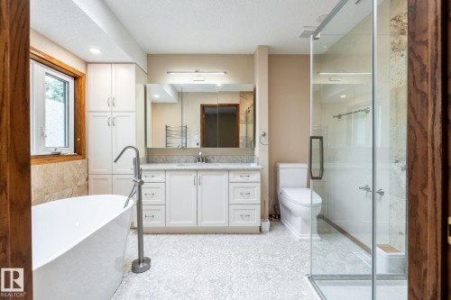 Contemporary en-suite bathroom featuring a freestanding tub with a floor-mounted faucet, a glass-enclosed shower, a double vanity with stone countertops, white shaker cabinetry, and pebble-tile flooring - 4632 151 Street, Edmonton, AB - Indoor Photo Showing Bathroom