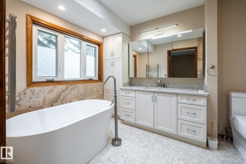 Freestanding soaking tub with a floor-mounted faucet, complemented by a large three-pane window with wood-finish trim and natural stone-look tile surround - 4632 151 Street, Edmonton, AB - Indoor Photo Showing Bathroom