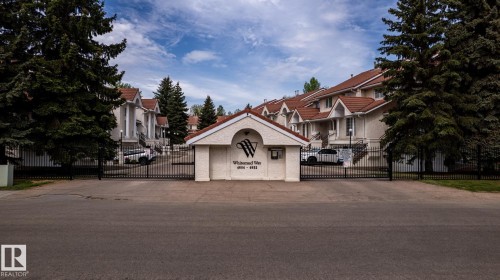 Gated community entrance featuring a stucco guardhouse with a tiled roof, wrought iron fencing, and mature evergreen trees - 4632 151 Street, Edmonton, AB - Outdoor With Facade