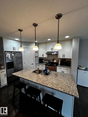 Kitchen featuring a spacious island with a speckled countertop, modern pendant lighting, white cabinetry, dark wood-finish flooring, and stainless steel appliances - 1868 30 Street Nw, Edmonton, AB - Indoor Photo Showing Kitchen With Upgraded Kitchen
