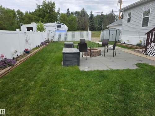 Expansive backyard featuring a stone-look paver patio, a built-in fire pit with stone-look surround, manicured lawn, and perimeter fencing - 5012 53 Ave, Breton, AB 