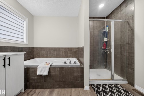 Bathroom featuring a built-in tub with tile surround, a glass-enclosed shower with tile walls, wood-finish flooring, a white vanity with dark hardware, and a window with horizontal blinds - 6085 King Landing, Edmonton, AB - Indoor Photo Showing Bathroom