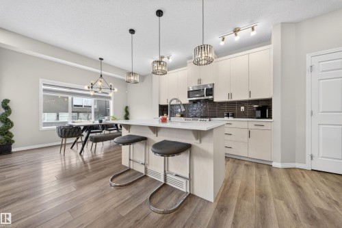 Open-concept living space featuring wood-finish flooring, a kitchen island with seating, pendant lighting fixtures, light-toned cabinetry, and a dark tile backsplash - 6085 King Landing, Edmonton, AB - Indoor Photo Showing Kitchen With Upgraded Kitchen