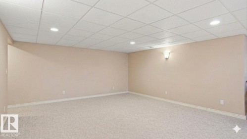 Spacious room featuring neutral carpeting, light beige walls, a drop ceiling with recessed lighting, and a wall sconce providing ambient illumination - 4309 37A Avenue, Edmonton, AB - Indoor