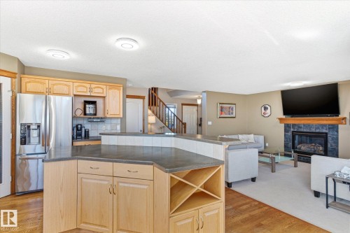 Open-concept kitchen featuring light wood cabinetry, stainless steel refrigerator, and a breakfast bar with a tile accent - 442 Hunters Green, Edmonton, AB - Indoor Photo Showing Kitchen With Fireplace