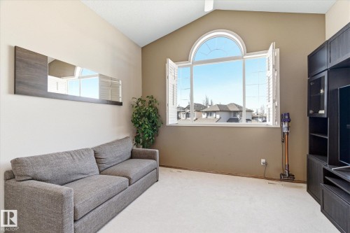 Vaulted ceiling room featuring a large arched window with white louvered shutters, light beige carpet flooring, and a built-in dark wood-finish media cabinet - 442 Hunters Green, Edmonton, AB - Indoor Photo Showing Other Room