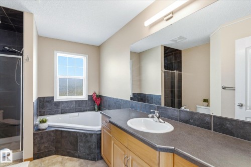 Corner soaking tub with a tiled surround, framed window, separate glass-enclosed shower with dark tile, and a vanity with a counter-mounted sink and light wood cabinetry - 442 Hunters Green, Edmonton, AB - Indoor Photo Showing Bathroom
