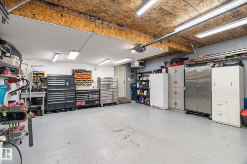 Garage space featuring a textured concrete floor, multiple fluorescent light fixtures, exposed wood-finish ceiling, a built-in workbench, and extensive storage solutions - 442 Hunters Green, Edmonton, AB - Indoor Photo Showing Garage