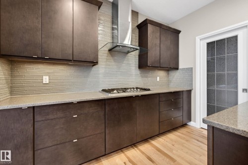 Contemporary kitchen featuring dark wood-finish cabinetry, light-toned countertops, a horizontal tile backsplash, stainless steel range hood, and light wood-finish flooring - 17838 60A Street, Edmonton, AB - Indoor Photo Showing Kitchen