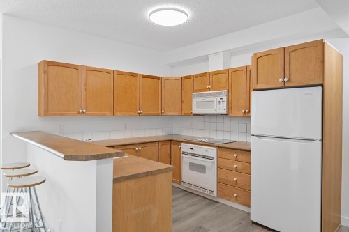 Kitchen featuring wood-finish cabinetry, white tile backsplash, a built-in microwave, integrated oven, and a breakfast bar - 117 10717 83 Avenue, Edmonton, AB - Indoor Photo Showing Kitchen