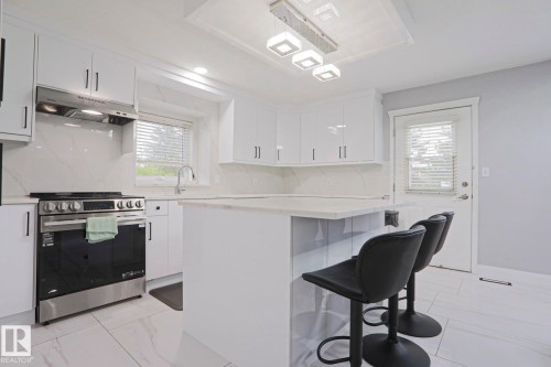 Bright kitchen featuring white cabinetry, a large kitchen island with counter seating, stainless steel appliances, a white tile backsplash, and light-toned tile flooring - 313 Jillings Crescent, Edmonton, AB - Indoor Photo Showing Kitchen