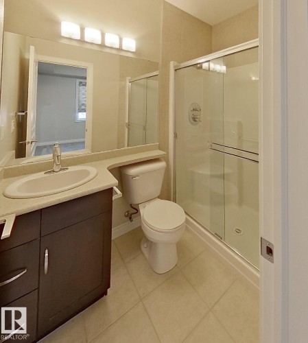Bathroom featuring a dark wood-finish vanity with an integrated sink, a rectangular mirror with four-bulb vanity lighting, a commode, and a glass-enclosed shower with tile surround - 108 9603 98 Avenue Nw, Edmonton, AB - Indoor Photo Showing Bathroom