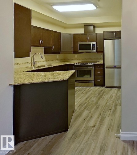Kitchen featuring wood-finish cabinetry, stainless steel appliances, granite-patterned countertops, a breakfast bar, and wood-look flooring - 108 9603 98 Avenue Nw, Edmonton, AB - Indoor Photo Showing Kitchen With Stainless Steel Kitchen