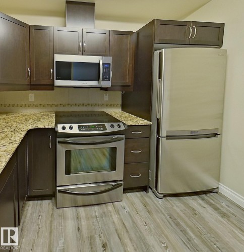 Kitchen featuring dark wood cabinetry, stainless steel appliances, granite countertops, a tiled backsplash, and wood-finish flooring - 108 9603 98 Avenue Nw, Edmonton, AB - Indoor Photo Showing Kitchen With Stainless Steel Kitchen