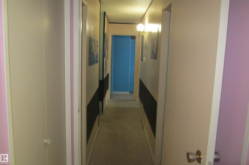 Hallway featuring textured carpet flooring, light beige wall paint, a blue door at the far end, and a wall-mounted light fixture - 3814 37 Avenue, Leduc, AB - Indoor