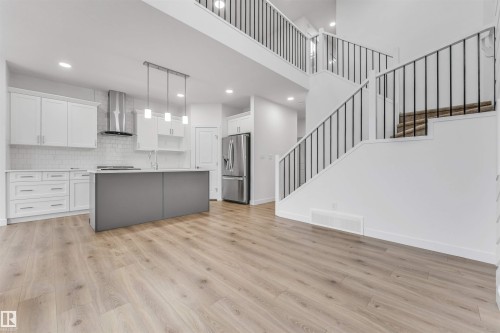 Open-concept living area featuring light wood-finish flooring, a contemporary kitchen with white cabinetry, a gray kitchen island, stainless steel appliances, and a two-story staircase with black metal railings - 1972 210 Street, Edmonton, AB - Indoor Photo Showing Kitchen With Upgraded Kitchen