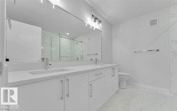Bathroom featuring a double vanity with white cabinetry and integrated sinks, a large wall-mounted mirror, overhead lighting, and a glass-enclosed shower with marble-look wall tiles - 