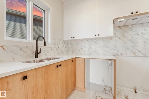 Kitchen featuring a dual-basin stainless steel sink with a matte black faucet, white upper cabinetry, light wood-finish lower cabinetry, and white countertops with a natural stone backsplash - 4015 39 Street, Beaumont, AB - Indoor Photo Showing Kitchen With Double Sink