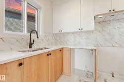 Kitchen featuring a dual-basin stainless steel sink with a matte black faucet, white upper cabinetry, light wood-finish lower cabinetry, and white countertops with a natural stone backsplash - 