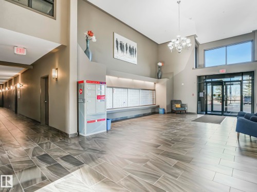 Lobby entrance featuring large format tile flooring, high ceilings, a contemporary chandelier, mailboxes, and a glass-paneled entry door with transom windows - 322 1520 Hammond Gate Nw, Edmonton, AB - Indoor Photo Showing Other Room