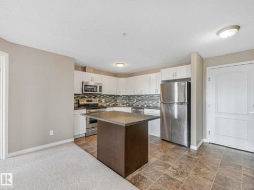 Kitchen featuring white cabinetry, stainless steel appliances, a multi-tone tile backsplash, and a central island with a dark countertop - 322 1520 Hammond Gate Nw, Edmonton, AB - Indoor Photo Showing Kitchen