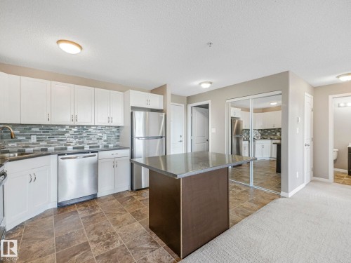 Kitchen featuring white cabinetry, stainless steel appliances, a tiled backsplash, and a central island with a dark countertop - 322 1520 Hammond Gate Nw, Edmonton, AB - Indoor Photo Showing Kitchen With Double Sink With Upgraded Kitchen