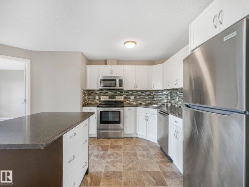 Kitchen featuring an island with a dark countertop, white cabinetry with chrome hardware, full suite of stainless steel appliances, and a mosaic tile backsplash - 322 1520 Hammond Gate Nw, Edmonton, AB - Indoor Photo Showing Kitchen With Upgraded Kitchen