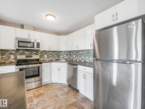 Kitchen featuring white cabinetry, stainless steel appliances, mosaic tile backsplash, dark countertops, and tile flooring - 322 1520 Hammond Gate Nw, Edmonton, AB - Indoor Photo Showing Kitchen With Upgraded Kitchen