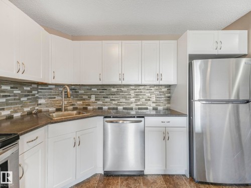 Kitchen featuring white cabinetry, dark countertops, a mosaic tile backsplash, stainless steel appliances, and wood-finish flooring - 322 1520 Hammond Gate Nw, Edmonton, AB - Indoor Photo Showing Kitchen With Upgraded Kitchen