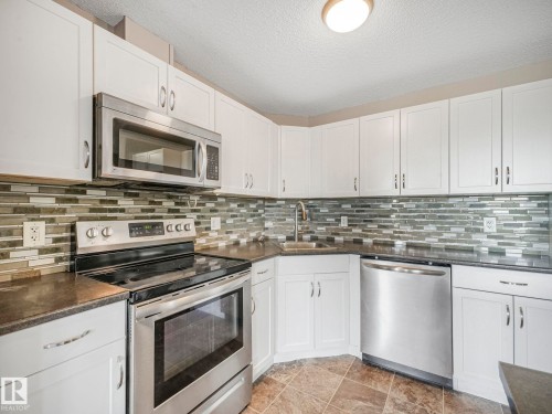 Modern kitchen featuring white cabinetry, stainless steel appliances, dark countertops, a mosaic tile backsplash, and tile flooring - 322 1520 Hammond Gate Nw, Edmonton, AB - Indoor Photo Showing Kitchen With Upgraded Kitchen