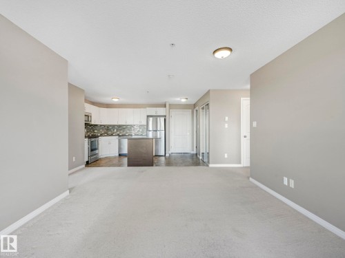 Open-concept living area with light-colored carpeting, featuring a kitchen with white cabinetry, stainless steel appliances, a dark-toned island with an overhang, and a tiled backsplash - 322 1520 Hammond Gate Nw, Edmonton, AB - Indoor