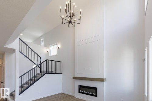 Double-height living space featuring a linear electric fireplace with a wood-finish mantel, decorative wall paneling, and a contemporary chandelier - 6275 180 Avenue Nw, Edmonton, AB - Indoor Photo Showing Other Room With Fireplace