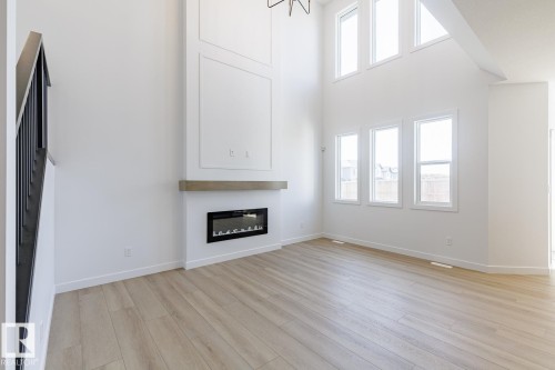 Two-story living area featuring a modern fireplace with a wood-finish mantel, wood-finish flooring, white walls, and multiple windows - 6275 180 Avenue Nw, Edmonton, AB - Indoor Photo Showing Living Room With Fireplace