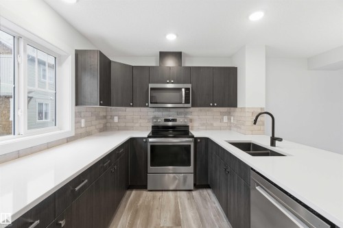 U-shaped kitchen featuring dark wood-finish cabinetry, light-toned brick-patterned backsplash, stainless steel appliances, a double basin sink with a matte black faucet, and light wood-finish flooring - 36 1005 Graydon Hill Boulevard, Edmonton, AB 