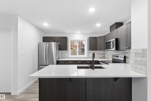 Kitchen featuring a large central island with a white countertop, dark wood-finish cabinetry, stainless steel appliances, a grey brick-patterned backsplash, and recessed lighting - 36 1005 Graydon Hill Boulevard, Edmonton, AB 