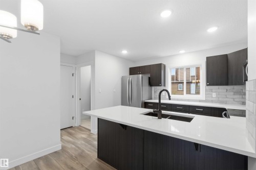Modern kitchen featuring a large island with a white countertop and integrated dual-basin sink, dark wood-finish cabinetry, stainless steel appliances, and a light-toned brick-patterned backsplash - 36 1005 Graydon Hill Boulevard, Edmonton, AB 