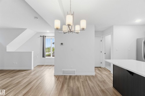 Open-concept living space featuring wood-finish flooring, a contemporary chandelier, a window with outdoor scenery, a kitchen island with a light countertop, and a stainless steel refrigerator - 36 1005 Graydon Hill Boulevard, Edmonton, AB 