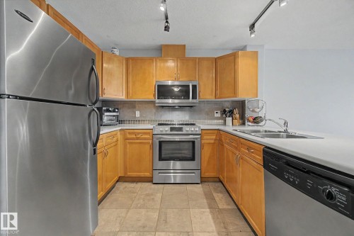 U-shaped kitchen featuring wood cabinetry, stainless steel appliances, light-toned countertops, and a tiled backsplash - 303 530 Hooke Road, Edmonton, AB - Indoor Photo Showing Kitchen With Double Sink