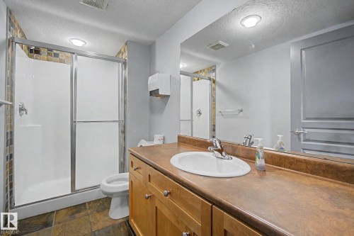 Full bathroom featuring a shower with sliding glass doors, a vanity with a brown countertop and integrated sink, light wood cabinetry, and stone tile flooring - 303 530 Hooke Road, Edmonton, AB - Indoor Photo Showing Bathroom