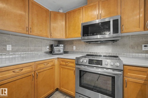 Kitchen featuring light wood cabinetry, a stainless steel electric range, an over-the-range microwave, light-toned countertops, and a gray tile backsplash with a decorative accent border - 303 530 Hooke Road, Edmonton, AB - Indoor Photo Showing Kitchen